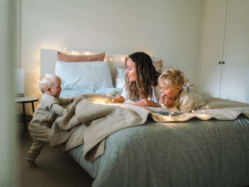 Mum and son lying on their belly and toddler baby climbing on the bed on the bed with string lights - Australian Stock Image