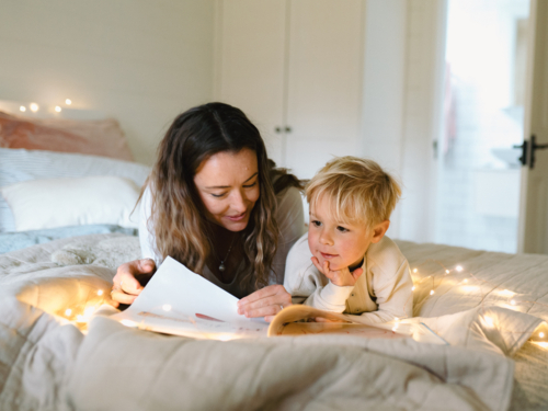 Mum and son lying on their belly on the bed with string lights reading a book - Australian Stock Image