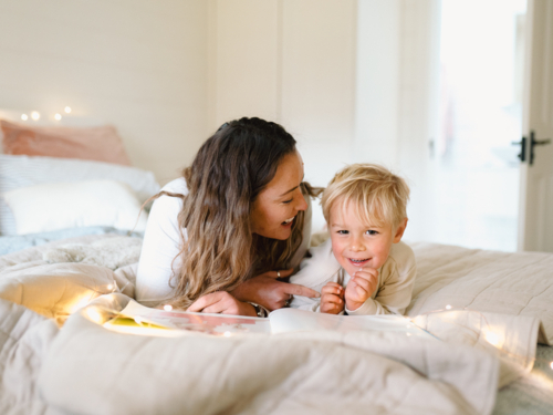 Mum and toddler son lying on their belly on the bed with string lights reading a book - Australian Stock Image