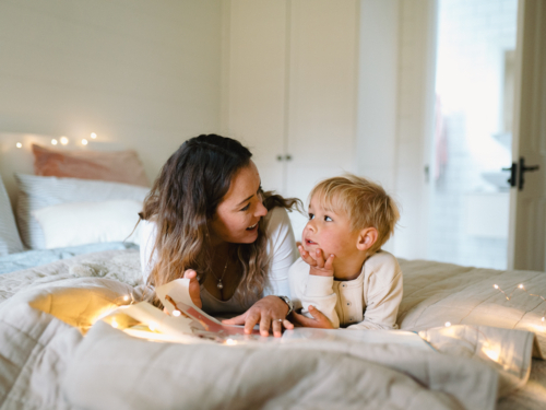 Mum and toddler son lying on their belly on the bed with string lights reading a book - Australian Stock Image