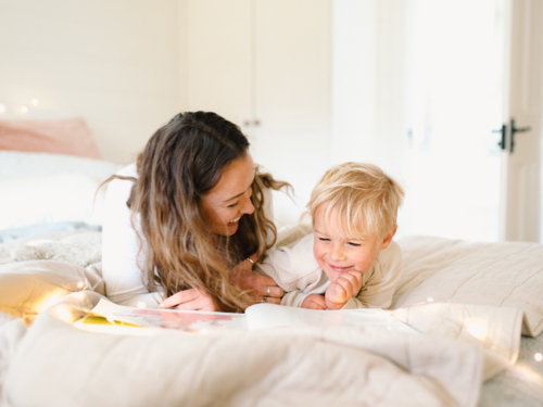 Mum and toddler son lying on their belly on the bed with string lights reading a book - Australian Stock Image