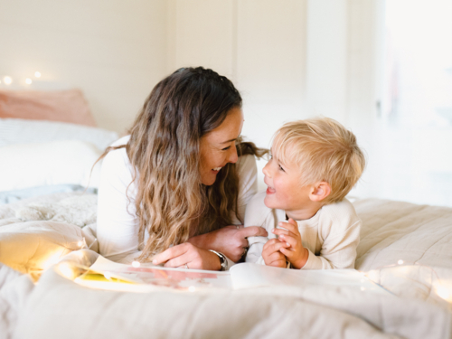 Mum and toddler son lying on their belly on the bed with string lights reading a book - Australian Stock Image