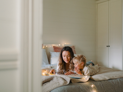 Mum and toddler son lying on their belly on the bed with string lights reading a book - Australian Stock Image