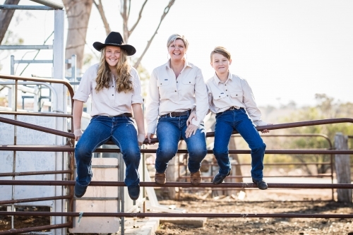 Mum and two children sitting on stock yards gate smiling on farm in drought - Australian Stock Image