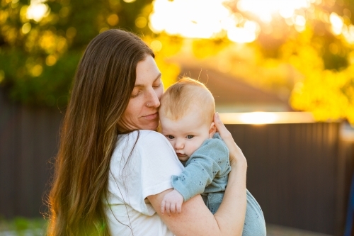 mum holding four month old baby girl in arms outside in golden bokeh sunset light in backyard - Australian Stock Image