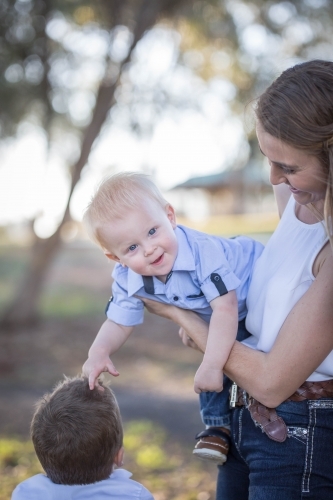 Mum holding laughing baby boy on hip while he plays with brother's hair - Australian Stock Image