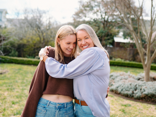 Mum hugging teenage daughter while standing in the backyard of their new home on a sunny day - Australian Stock Image