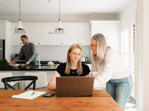 Mum leaning over teenage daughter working on her laptop on the wooden table - Australian Stock Image