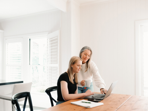 Mum leaning over teenage daughter working on her laptop on the wooden table - Australian Stock Image