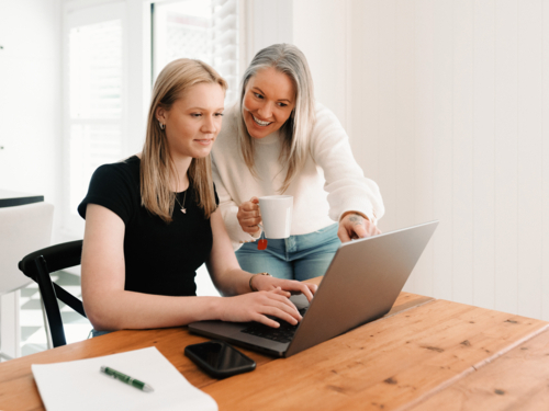 Mum leaning over teenage daughter working on her laptop on the wooden table - Australian Stock Image