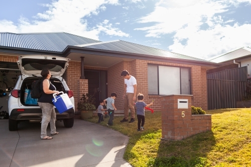 Mum packing family car before sports training with kids playing with hockey sticks on front lawn - Australian Stock Image