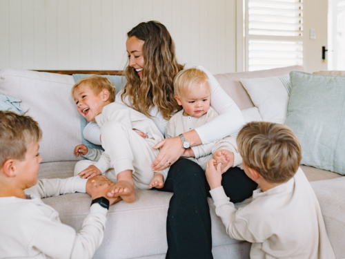 Mum sitting on the couch cradling her two sons while older brothers tickling their feet - Australian Stock Image
