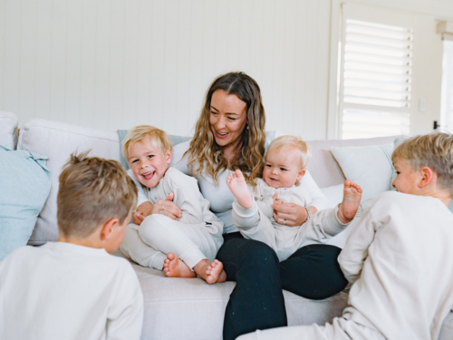 Mum sitting on the couch cradling her two sons while the other kneels on the floor - Australian Stock Image