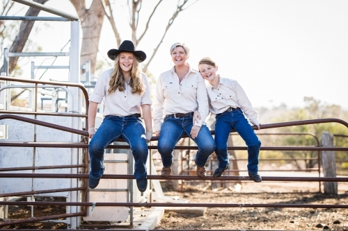 Mum sitting with children on fence in yards on farm in drought - Australian Stock Image