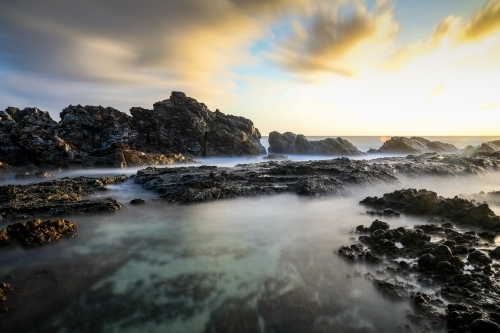 Ocean and rocky coastline against cloudy sunrise - Australian Stock Image