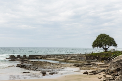 Ocean pool on beach with rocks and sand and tree with cloudy skies - Australian Stock Image