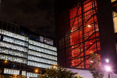 Office building at night, windows illuminated white and stairwell of other building illuminated red