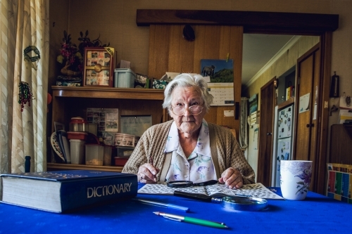 Old lady at dining table with crossword, magnifying glass, dictionary and cup of tea