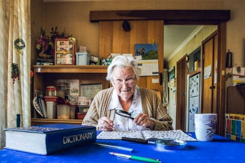 Old lady at dining table with crossword, magnifying glass, dictionary and cup of tea - Australian Stock Image