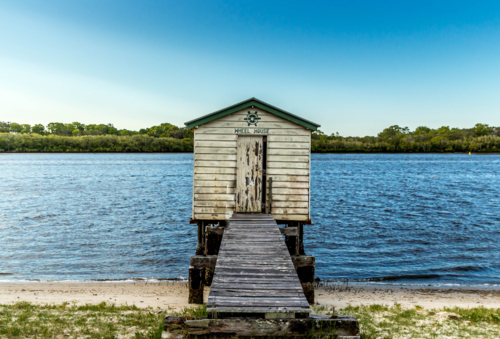 Old weathered wooden boat house with crooked jetty on river with blue sky and green vegetation - Australian Stock Image