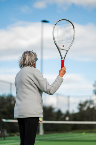 Older female tennis player ready to serve on a green court with a clear sky - Australian Stock Image