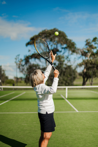 Older woman in white long-sleeve shirt serves tennis ball outdoors in daylight - Australian Stock Image