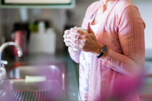 Older woman standing in kitchen holding a cuppa tea - Australian Stock Image
