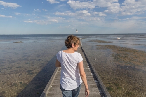 Older woman walking on timber pier looking over tidal shallows - Australian Stock Image