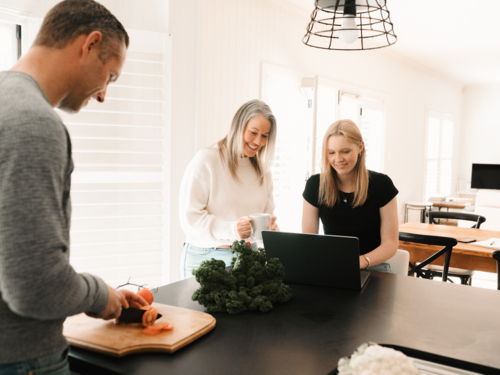 One male adult preparing food while mum helps teen daughter with work on laptop in bright kitchen - Australian Stock Image