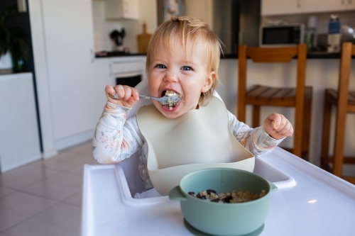 One year old baby girl using spoon to eat weetbix and sultanas for breakfast - Australian Stock Image