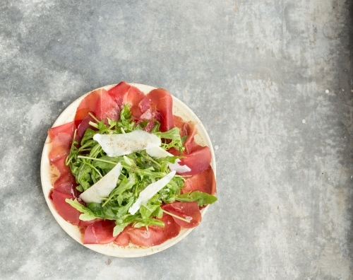 Overhead, minimalist image of bresaola, rocket and parmesan salad on a concrete table