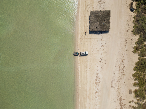 Overhead view of a  white vehicle and small boat sit at the edge of a sandy shoreline - Australian Stock Image