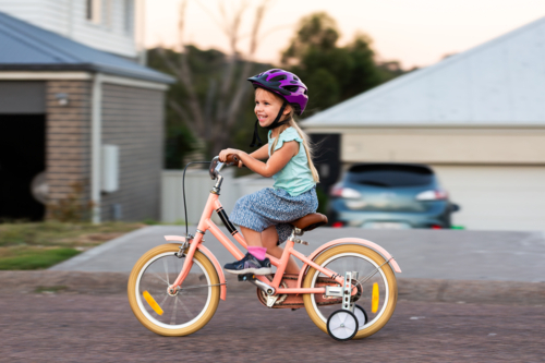 Panning movement shot of preschooler riding pink bike with training wheels on roadside - Australian Stock Image