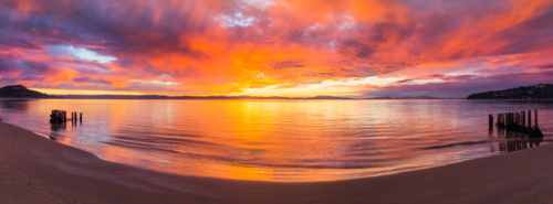 Panorama view of a colourful sunrise reflected in a calm ocean bay with wooden groynes - Australian Stock Image
