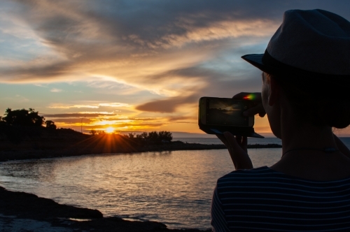 Partial silhouette of woman taking photo of sunset on mobile phone - Australian Stock Image