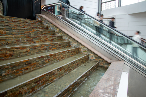 People on an escalator in a shopping centre with a water feature in the foreground - Australian Stock Image