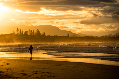 Person walking along a beach at sunset with golden light and distant mountains - Australian Stock Image