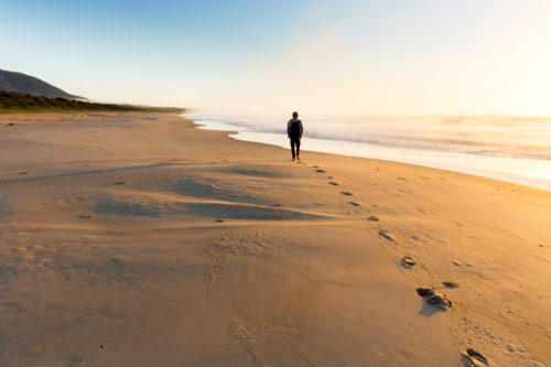 Person walking on a smooth beach leaves a trail of footprints in the sand during golden sunrise - Australian Stock Image