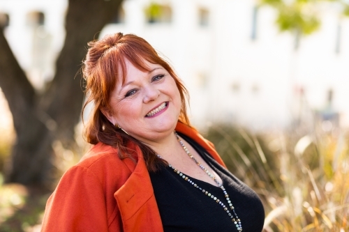portrait of a happy middle aged redhead person outside with orange jacket - Australian Stock Image