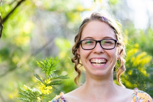 Portrait of a happy smiling young woman with glasses standing among leaves and blossoms of wattle - Australian Stock Image