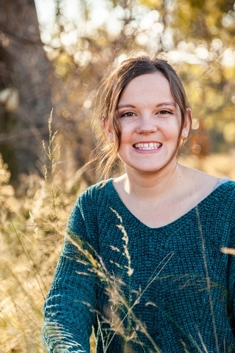 Portrait of a happy young adult in green jumper siting in backlit grass - Australian Stock Image
