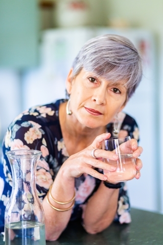 Portrait of a healthy senior woman leaning on the kitchen bench drinking a glass of water - Australian Stock Image