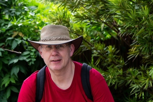 Portrait of a mature male tourist wearing an Australian hat and a backpack with greenery behind - Australian Stock Image