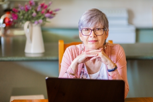 Portrait of a senior woman wearing glasses with hands under her chin sitting inside her home - Australian Stock Image