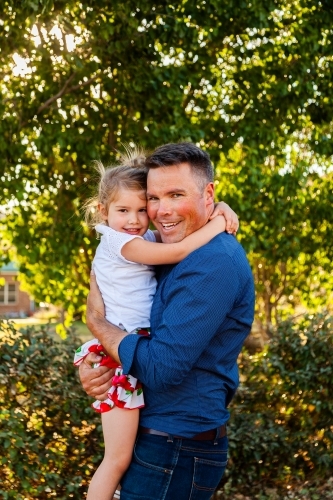 Portrait of a smiling father hugging his happy daughter in golden sunlight - Australian Stock Image