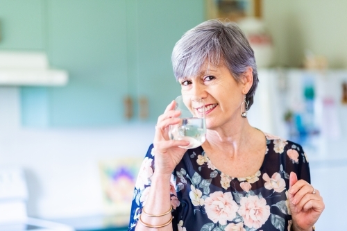 Portrait of a smiling woman drinking a glass of water in the kitchen - Australian Stock Image