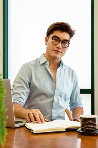 Portrait of a young male office worker sitting with laptop and note book at a meeting table - Australian Stock Image