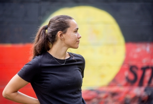 Portrait of an Aboriginal woman standing with a red, yellow and black flag mural in the background - Australian Stock Image