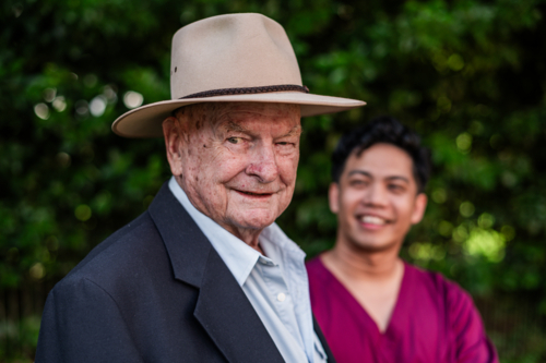 Portrait of an elderly man in a hat smiling and standing outdoors with male healthcare worker - Australian Stock Image