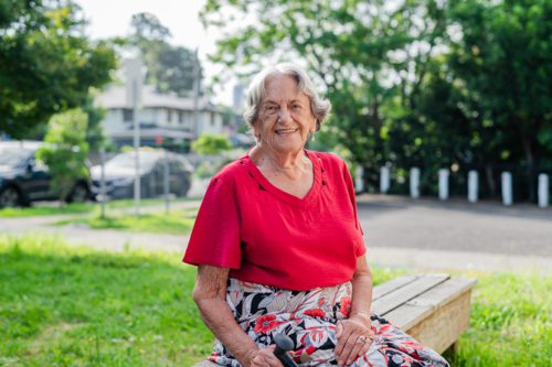 Portrait of an elderly woman in red blouse sitting on wooden bench outdoors smiling - Australian Stock Image